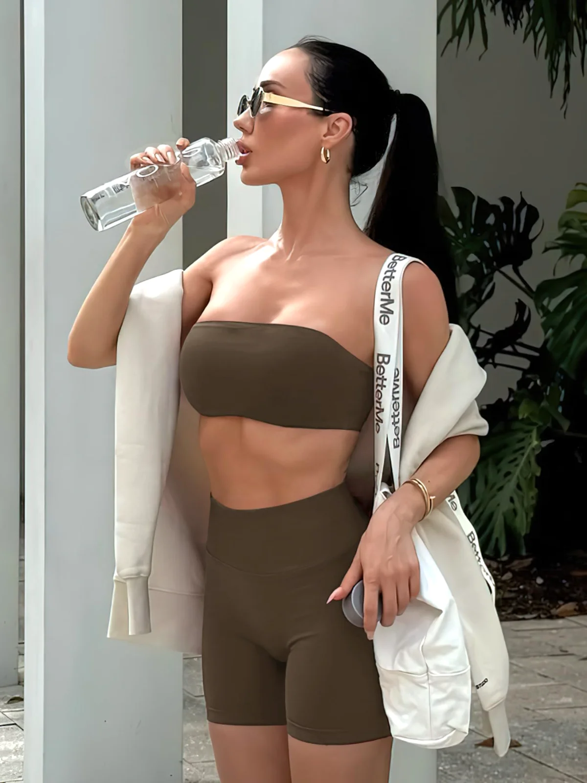 A coffee-brown bandeau bra and matching shorts on a sunlit street, drinking from a bottle, a bag on the arm